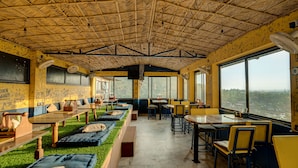 A restaurant with a thatched roof, green artificial grass seating, and large windows offering a view. - Shalom Backpackers Rishikesh (Narendranagar)