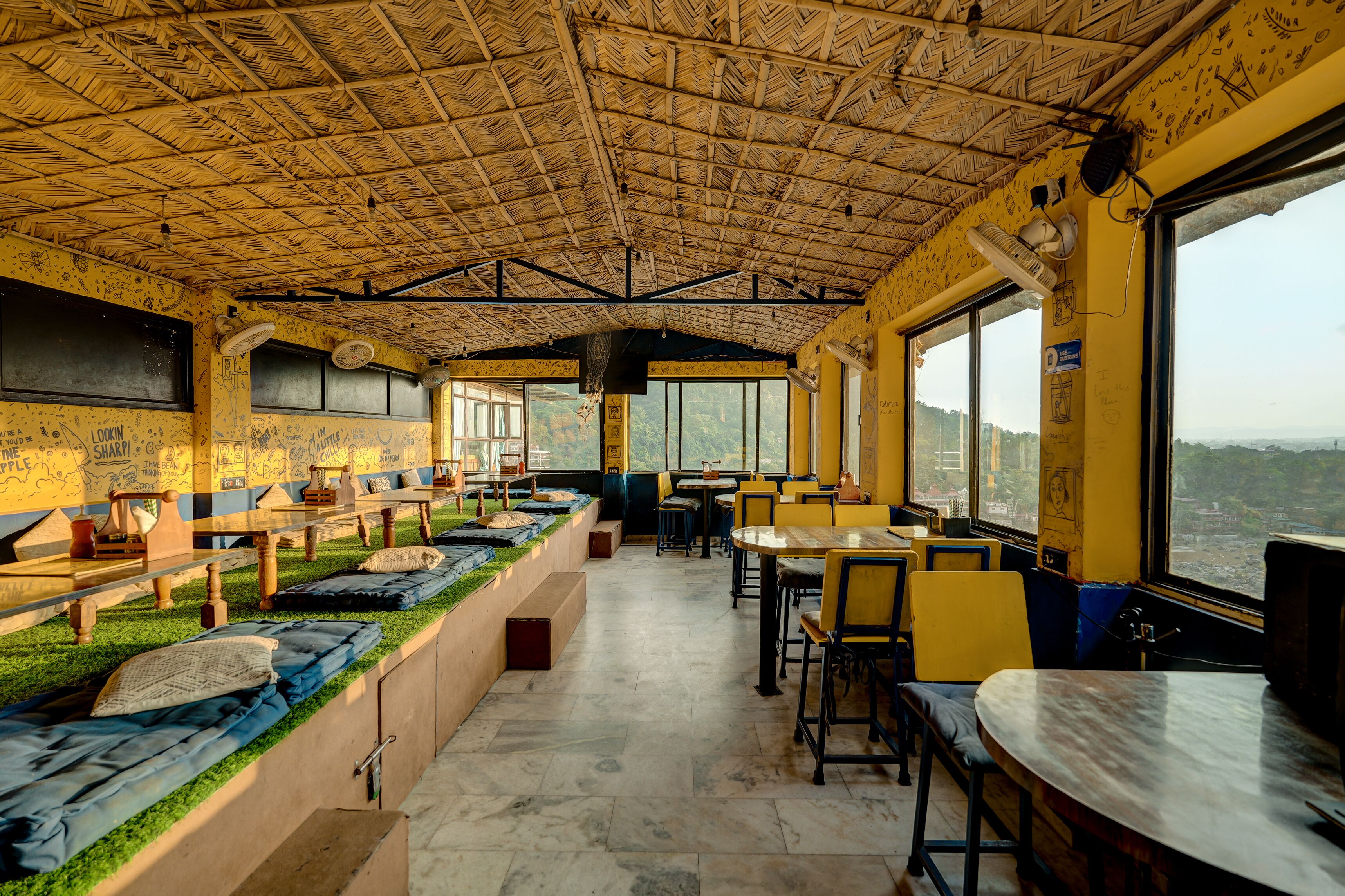 A restaurant with a thatched roof, outdoor seating with cushions, and large windows offering a view.