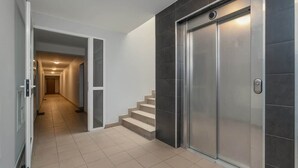A modern elevator with a metallic finish, a hallway with beige tiles, and a staircase leading to another floor.