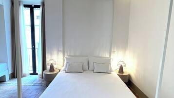 Una stanza con soffitto a mattonelle, un letto bianco con due cuscini, un comodino con una lampada e una porta che dà sull'esterno.