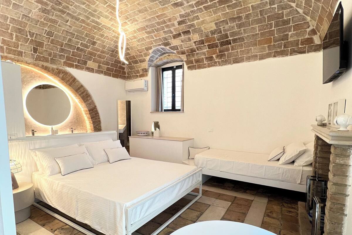 A bedroom with a brick ceiling, a round mirror, a bed, a bedside table, and a small window.
