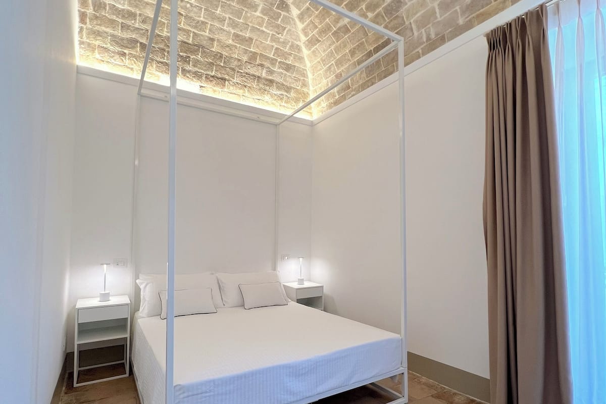 A bedroom with a four-poster bed, white walls, a brick ceiling, and a window with curtains.