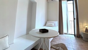 A small, modern room with a white bed, a round table with a black object, two woven chairs, a white bench, and a brick wall. - RESIDENZA ORTENSIA (Petacciato)