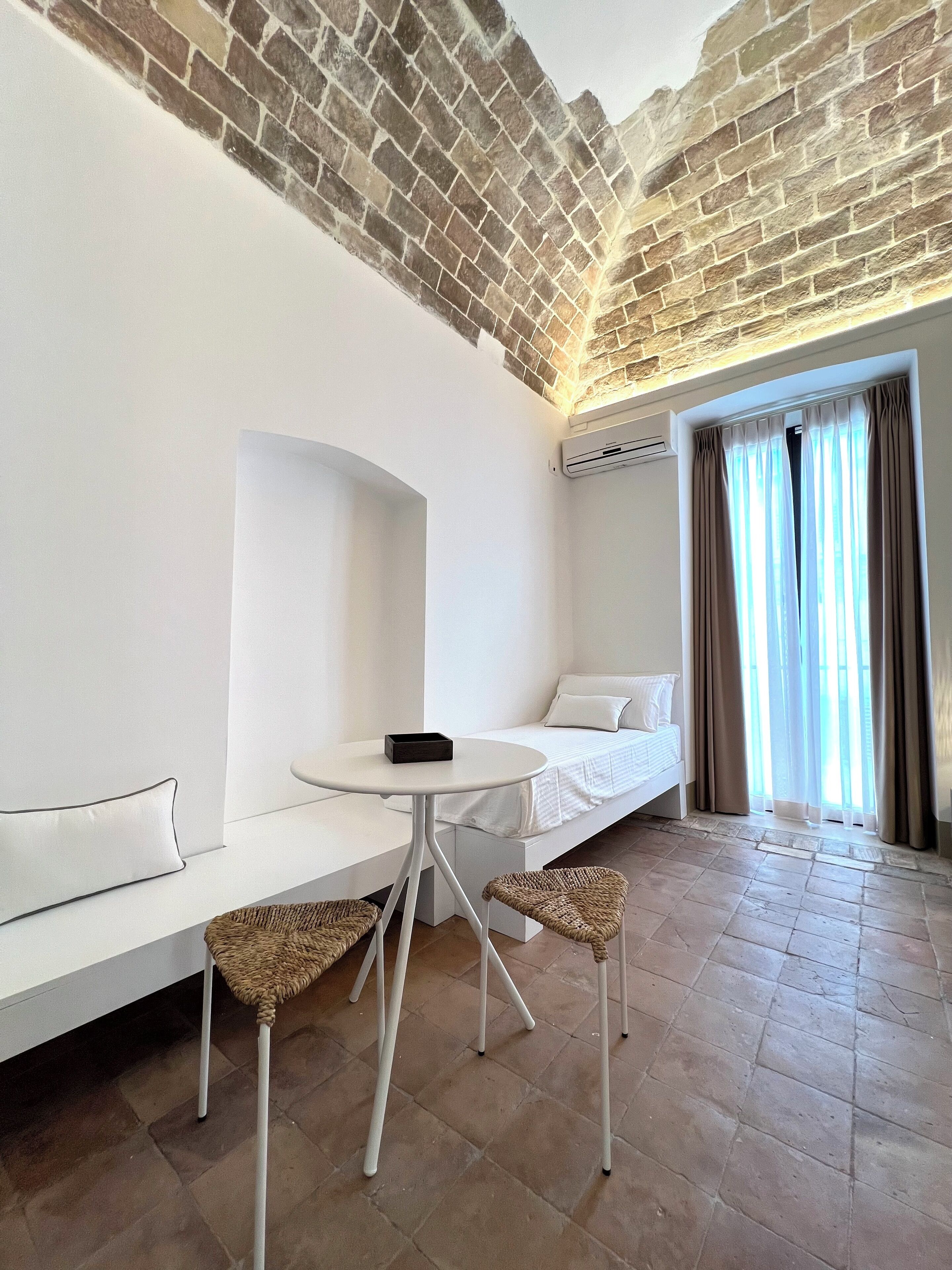 Una habitación moderna con techo de ladrillo, un sofá blanco, una mesita redonda pequeña, sillas de mimbre y una cama con ropa blanca de cama.