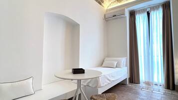Una habitación moderna con techo de ladrillo, un sofá blanco, una mesita redonda pequeña, sillas de mimbre y una cama con ropa blanca de cama.