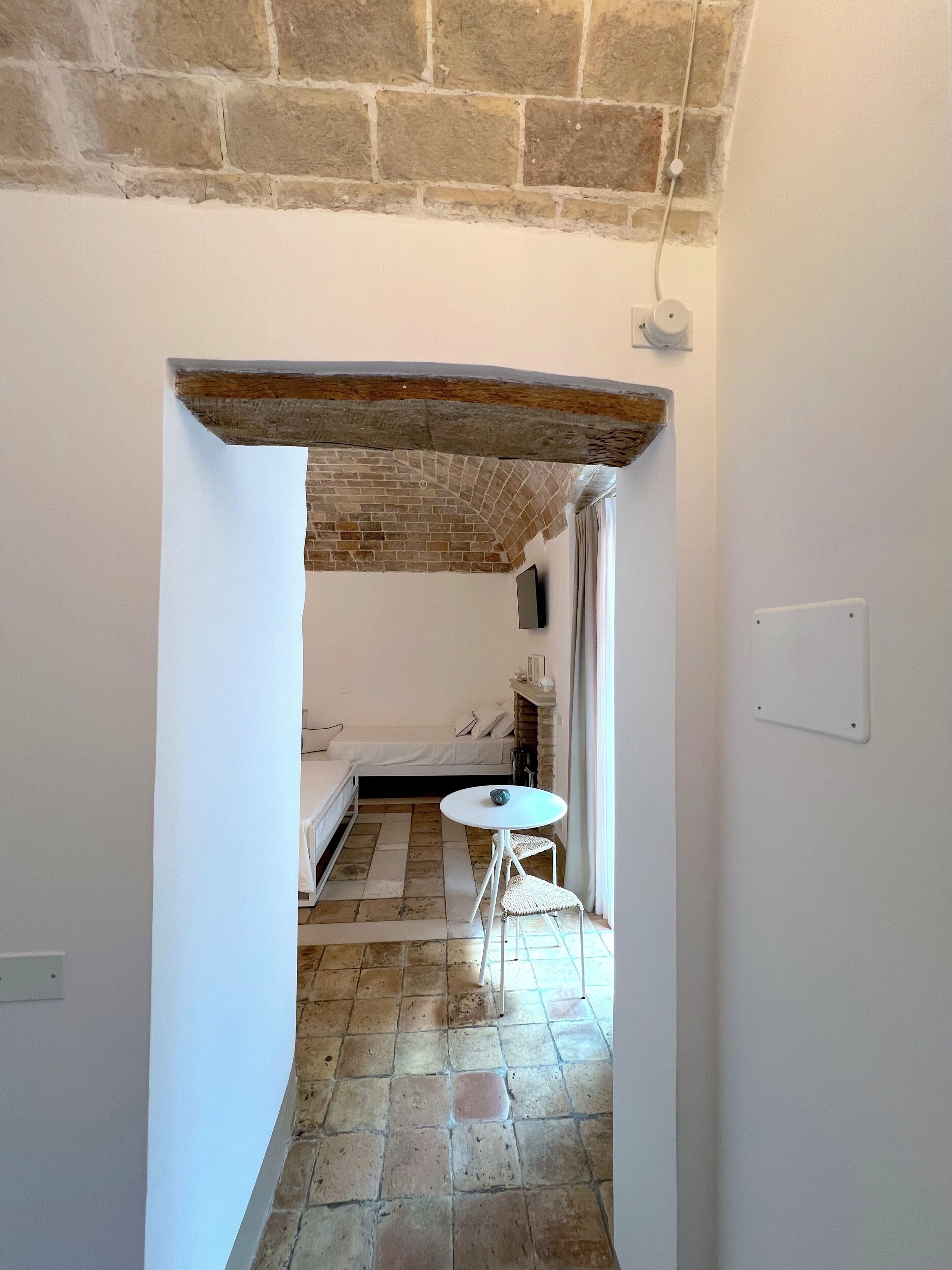 Un couloir étroit, doté d’un sol en pierre, mène à une chambre à coucher qui contient un lit, une petite table et un téléviseur.