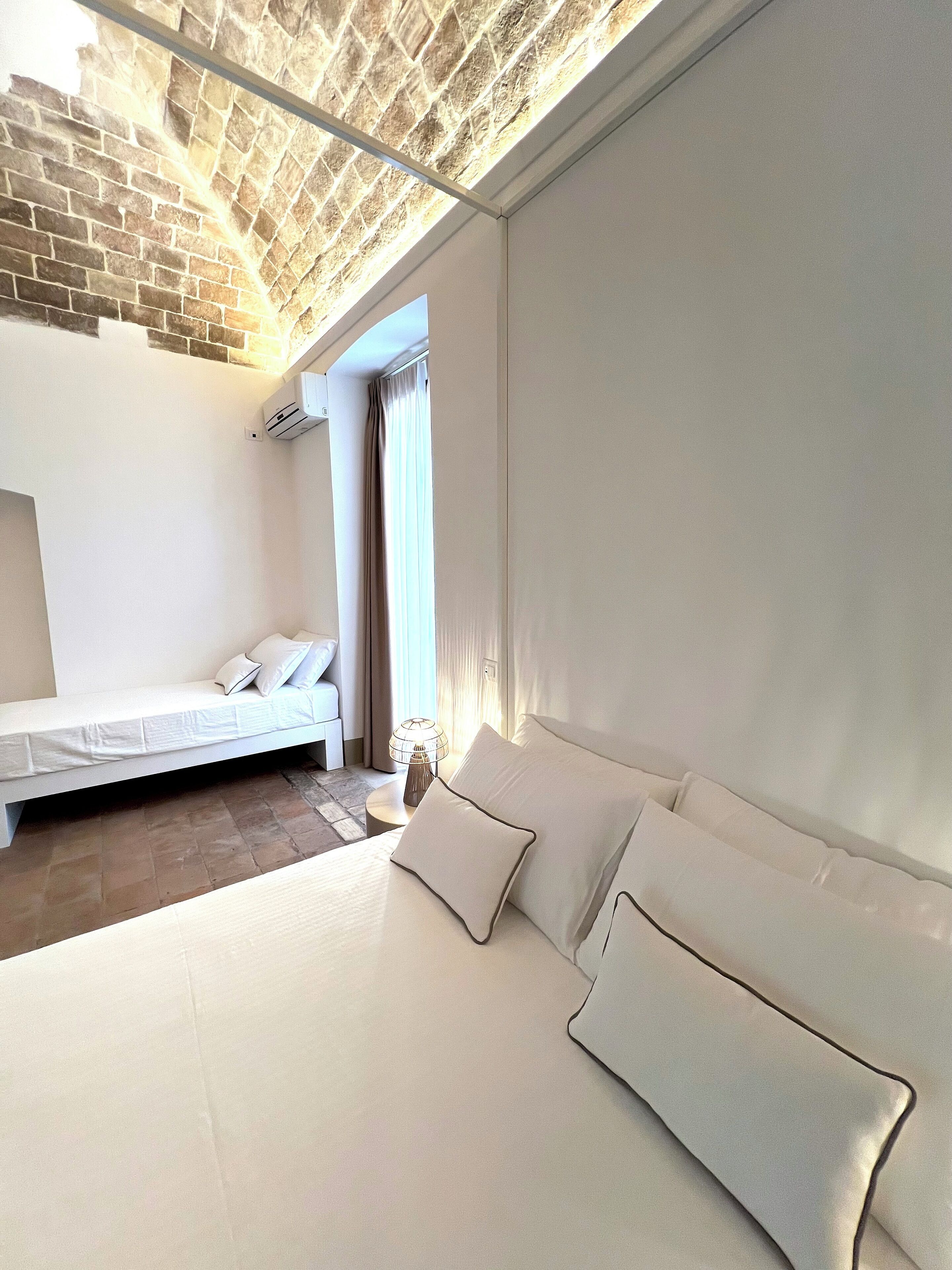A modern bedroom with a brick ceiling, a bed with white linens, a bedside table with a lamp, and a view of a cityscape through a window.