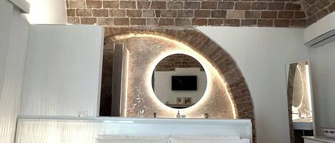 Een moderne slaapkamer met een bakstenen plafond, een ronde spiegel, een bed met witte lakens en een nachtkastje met een lamp.