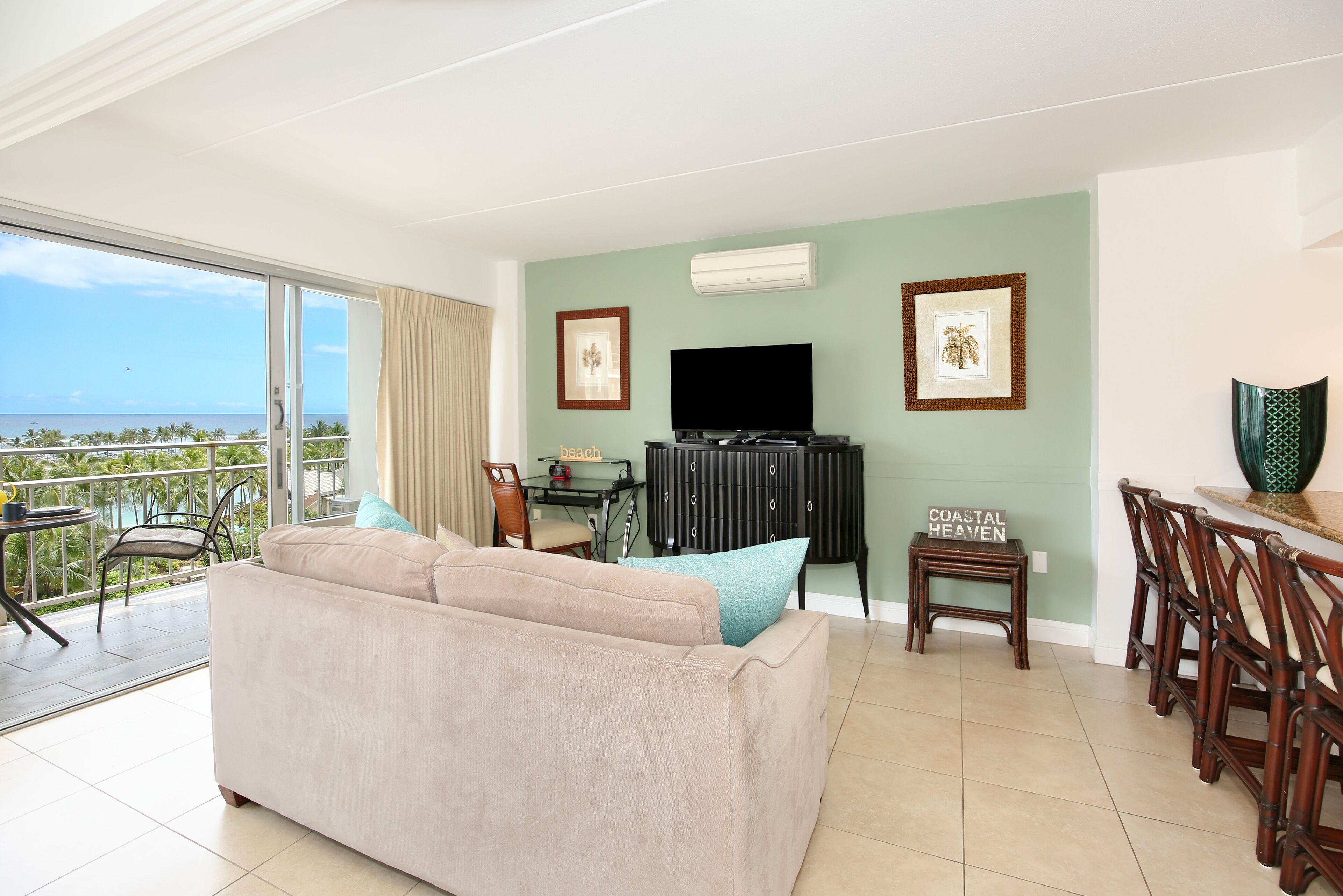 A living room with a sofa, a TV, a dining table, and chairs. There is a balcony with a view of the ocean.