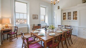 Uma sala de jantar com uma mesa de madeira posta para seis pessoas, rodeada por seis cadeiras com almofadas vermelhas. Há uma janela com vista para prédios, um radiador abaixo da janela e um candelabro sobre a mesa.