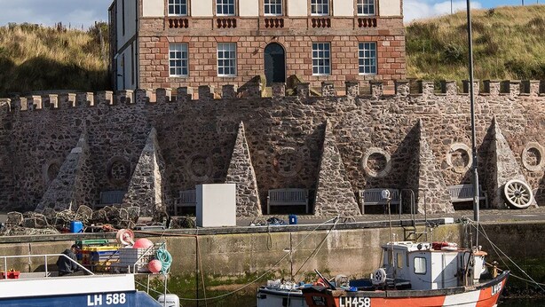 Um edifício histórico com parede de pedra e fosso, barcos atracados na frente, e céu claro.