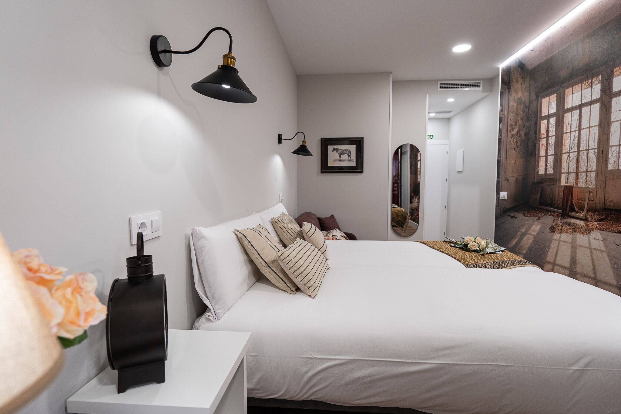 Ein modernes Hotelzimmer mit einem großen Bett, einem Nachttisch mit Lampe, einem gerahmten Bild an der Wand und einem Wandgemälde an der angrenzenden Wand.