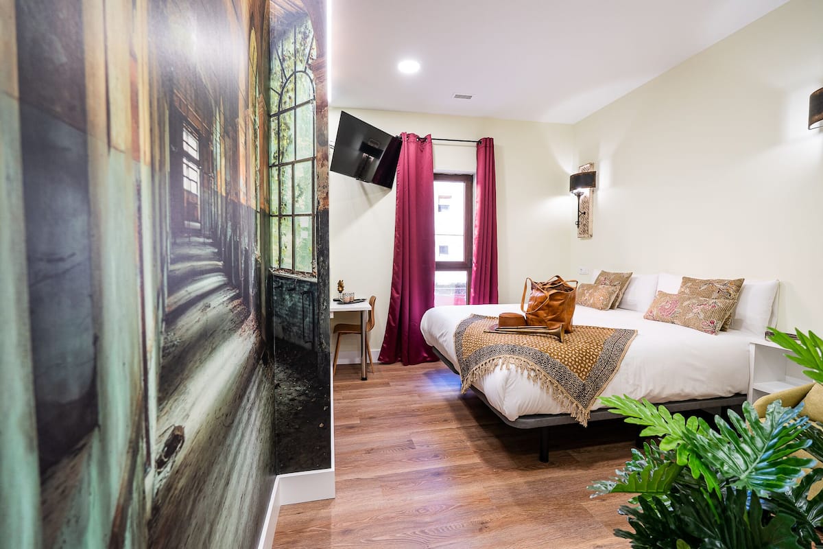 Une chambre à coucher avec un lit, un bureau, une télévision et une fenêtre avec des rideaux.