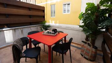 Un pequeño espacio al aire libre con comedor, una mesa roja y sillas negras, un banco de madera y una planta en maceta.