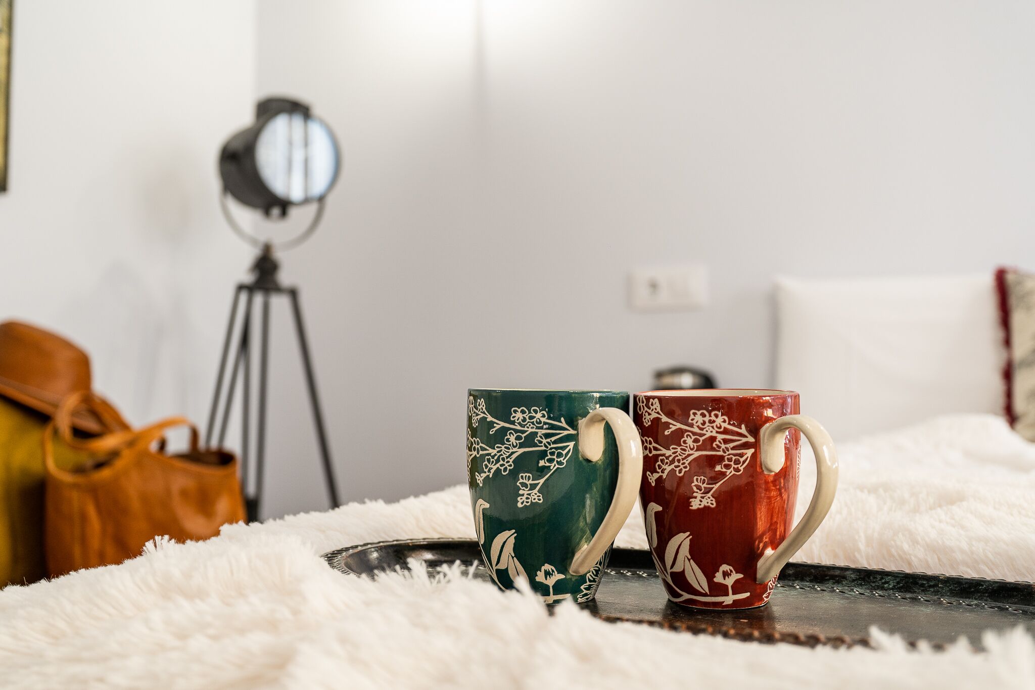 Two decorative mugs on a tray, a tripod, and a leather bag in a room with a white fluffy rug.