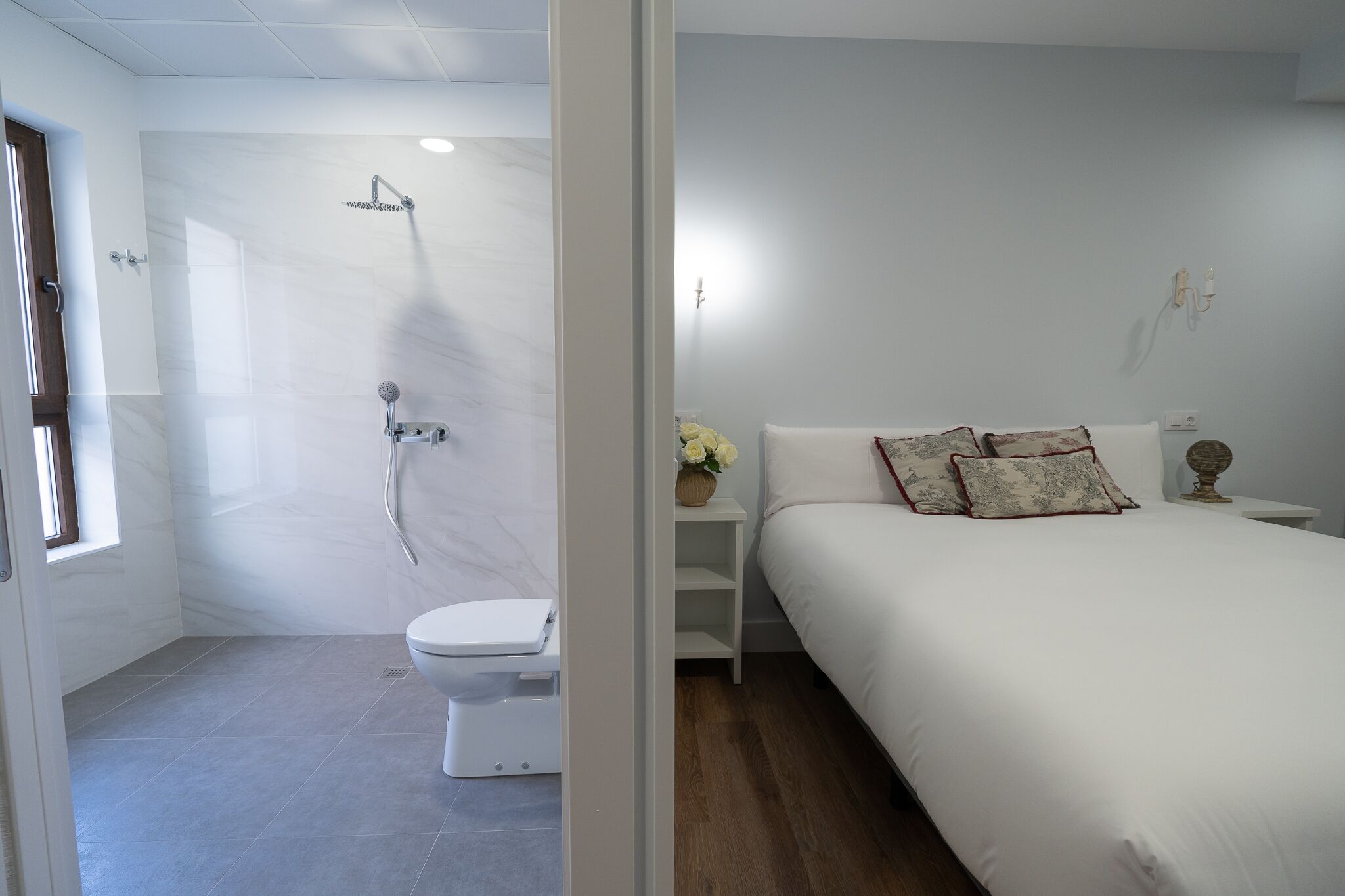 A modern bathroom with a shower, toilet, and a bed with white bedding and decorative pillows.