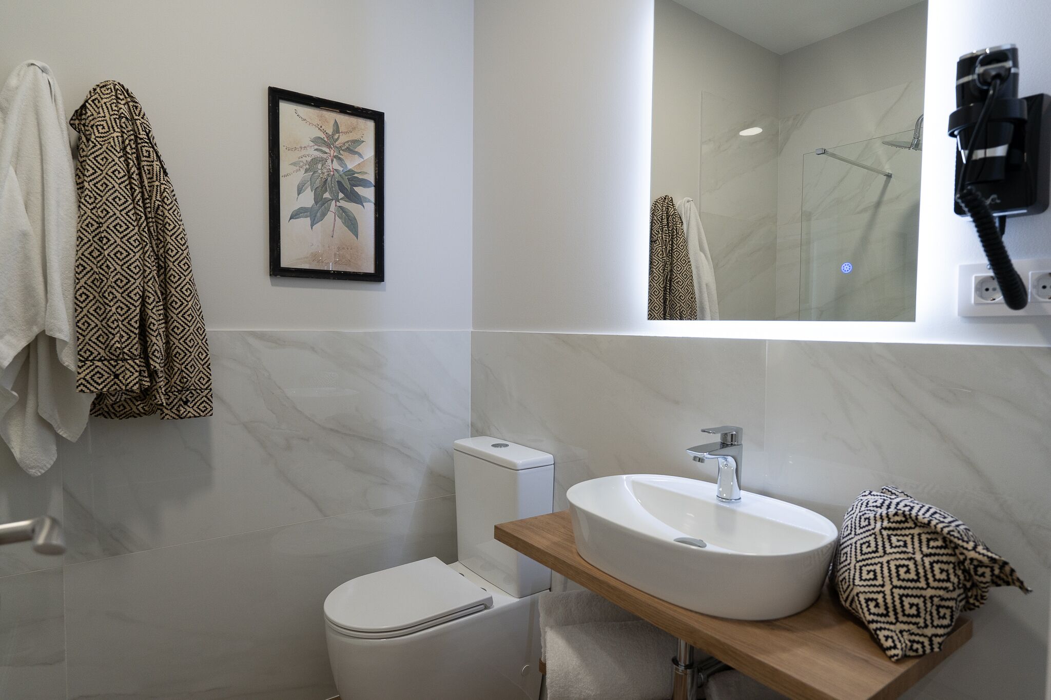 A modern bathroom with a white toilet, a white sink on a wooden countertop, a glass shower enclosure, and a towel rack with patterned towels.