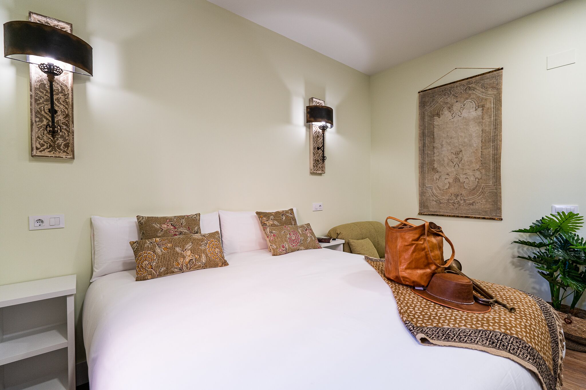 A neatly made bed with patterned pillows, a brown leather bag on the bed, and a large tapestry on the wall.