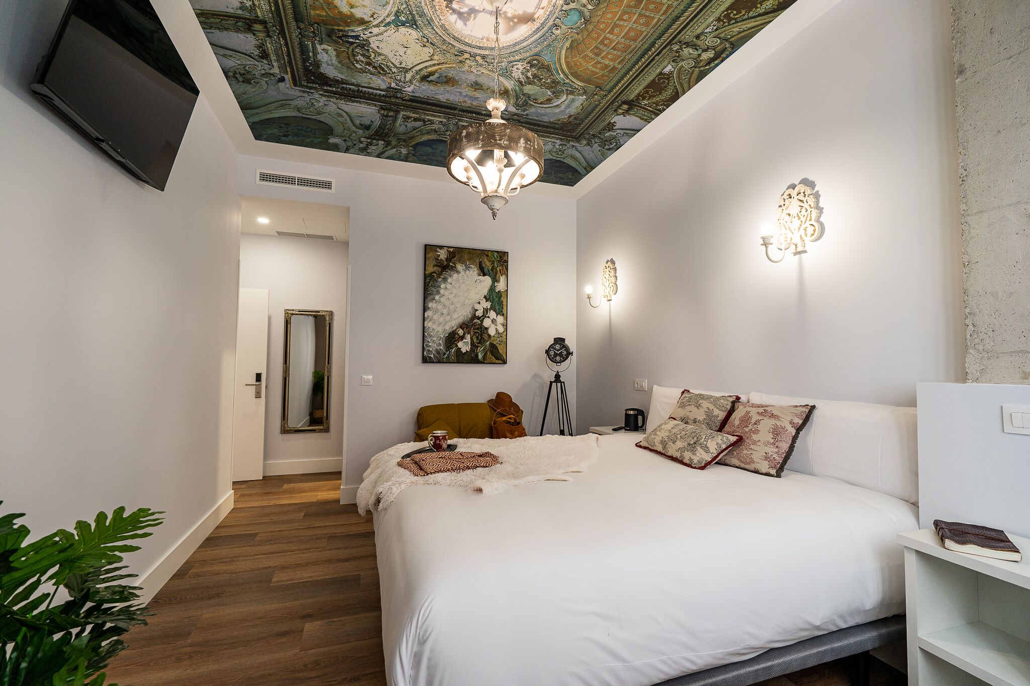 A bedroom with a large bed, a ceiling with a detailed fresco, a chandelier, and a mounted television.