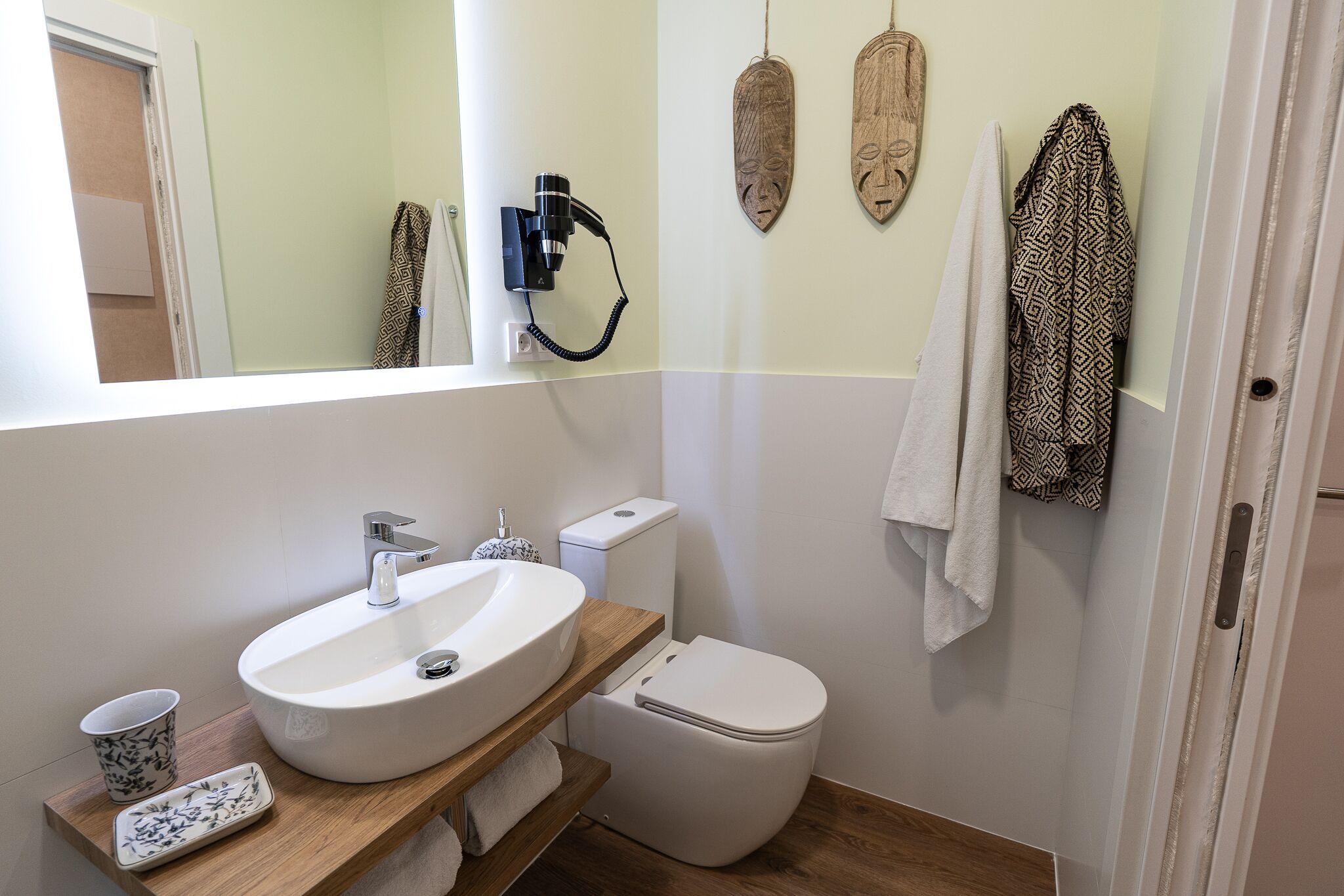 Un baño con lavamanos, inodoro y espejo blancos. Decoraciones de madera en la pared y una toalla colgada.