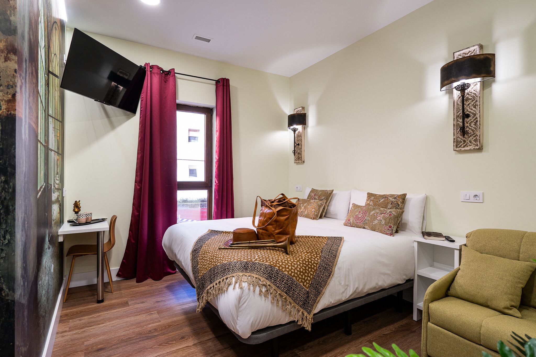 A hotel room with a bed, a desk, a chair, a TV mounted on the wall, and a window with curtains.
