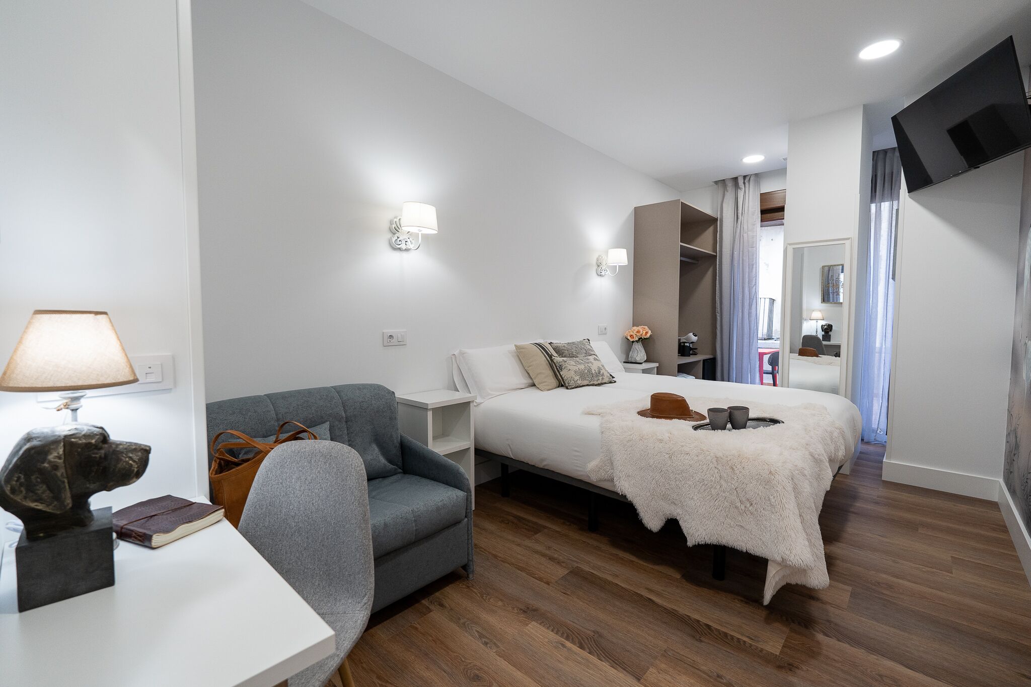 A hotel room with a bed, a chair, a desk with a lamp, and a TV mounted on the wall.