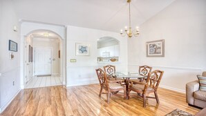 Un comedor con mesa de vidrio y sillas de madera, un candelabro y una cocina al fondo.