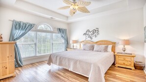 Un dormitorio con una cama grande, cabecera de madera, dos mesitas de noche con lámparas, un aparador de madera y una ventana con cortinas.