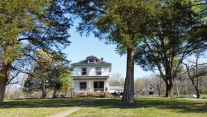 Une maison à deux étages avec un porche à l’avant, entourée d’arbres.