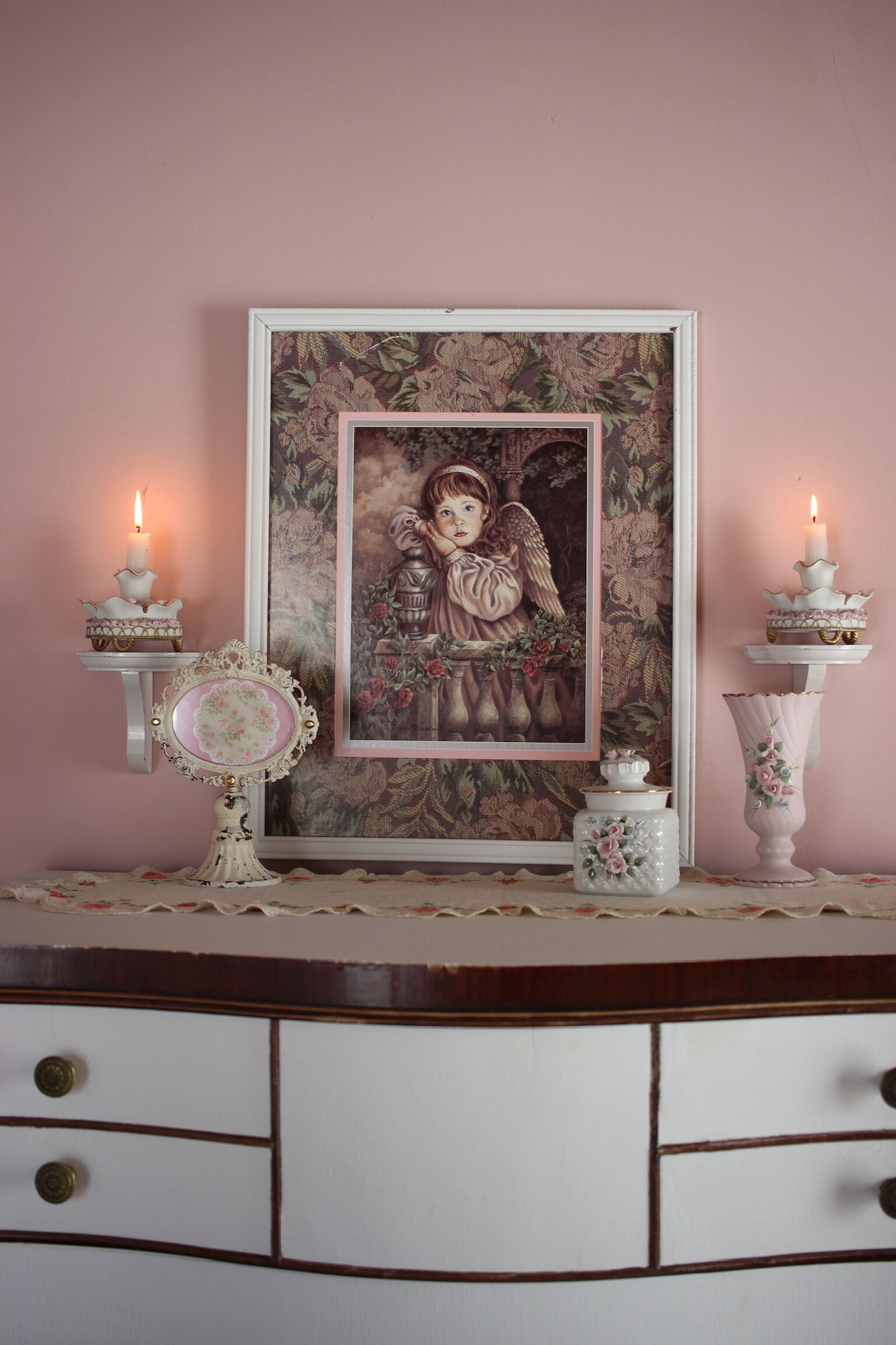 Une œuvre d’art encadrée accrochée au mur, deux bougies allumées sur des chandeliers, un miroir décoratif et une commode blanche avec des tiroirs.
