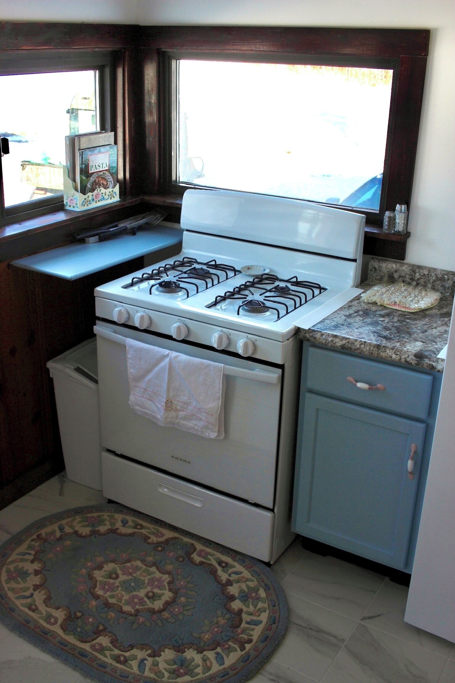 A kitchen with a gas stove, oven, and a small countertop. There is a window above the sink and a rug on the floor.