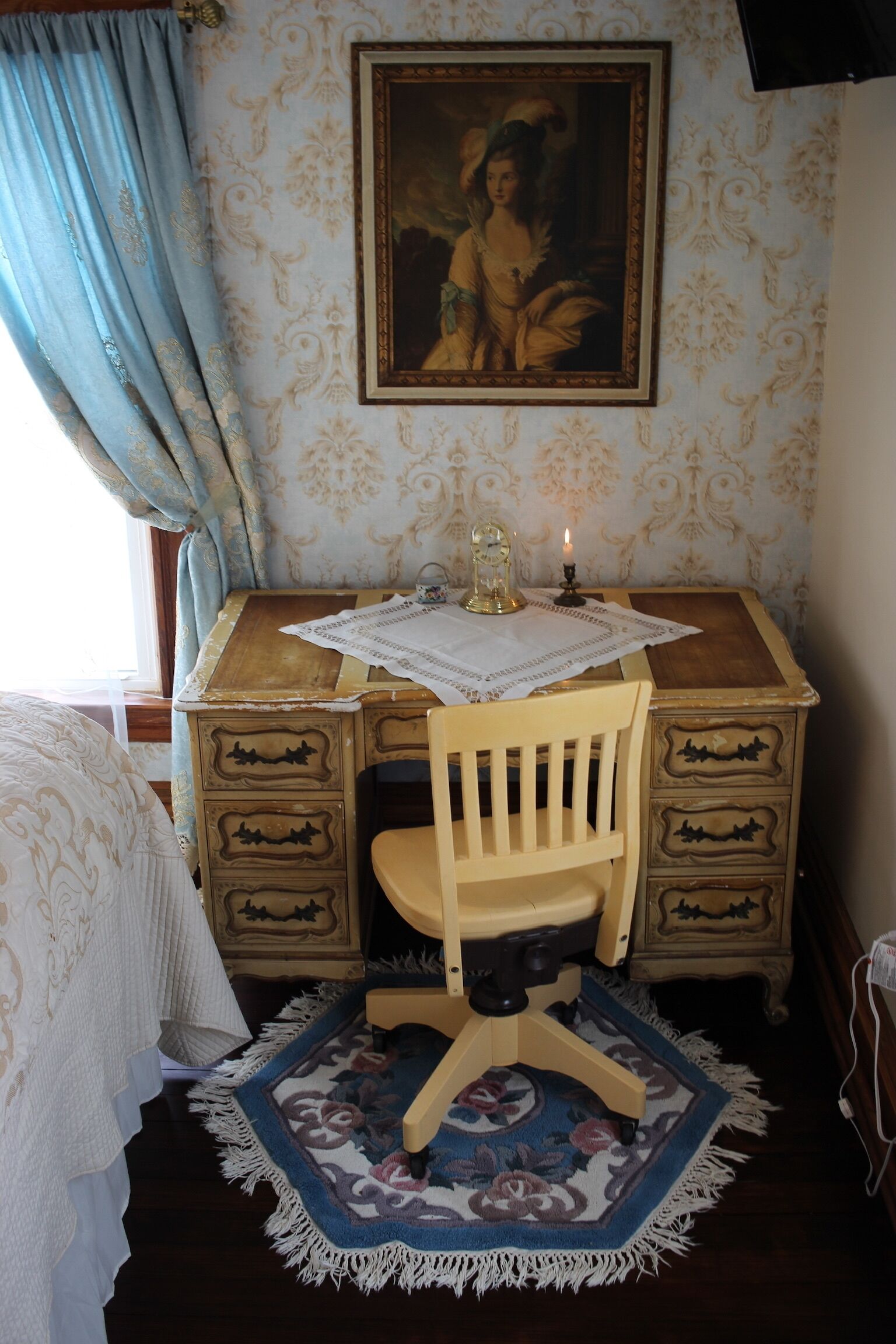 Una habitación con un escritorio de madera, una silla, una mesita con una vela, un cuadro enmarcado en la pared y una alfombra estampada en el suelo.