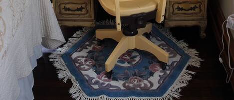 Una habitación con un escritorio de madera, una silla, una mesita con una vela, un cuadro enmarcado en la pared y una alfombra estampada en el suelo.
