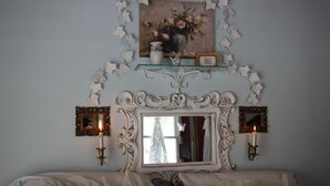 Une chambre à coucher avec un grand miroir décoratif, deux appliques murales, un tableau encadré au-dessus du miroir et une étagère décorative ornée d’un vase et d’un cadre photo.
