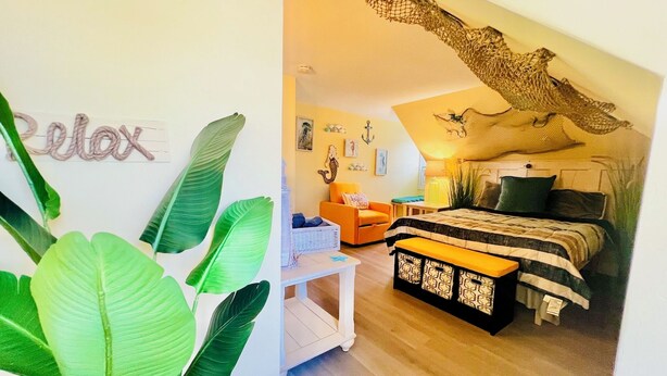 A cozy bedroom with a bed, a small table, and a chair. There are large green plants and a wall with a 'Relax' sign. - Cozy & Whimsical Nautical Theme Walk to Beach & Boardwalk (Wildwood)