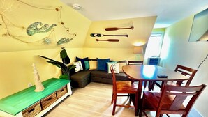 A cozy living room with a sofa, a dining table with chairs, a lamp, and a wall-mounted shelf with decorative items. - Cozy & Whimsical Nautical Theme Walk to Beach & Boardwalk (Wildwood)
