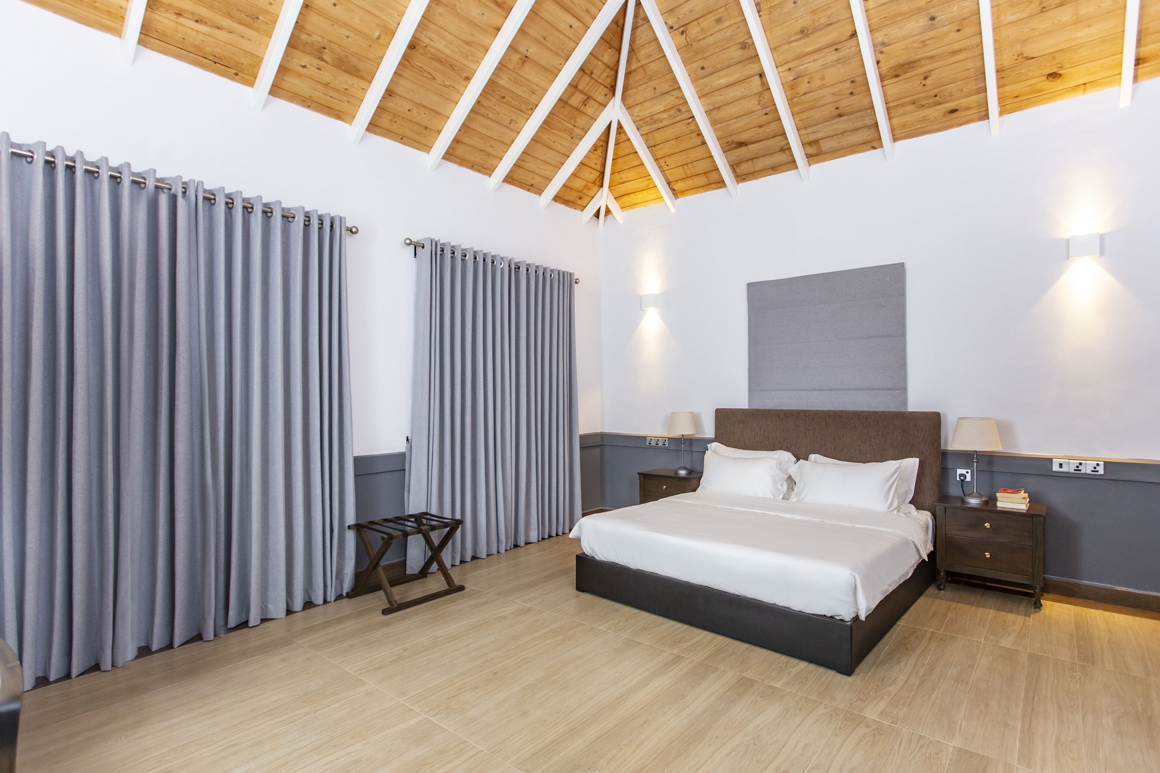 A modern hotel room with a wooden ceiling, a large bed, grey curtains, a bedside table with a lamp, and a wall-mounted light.