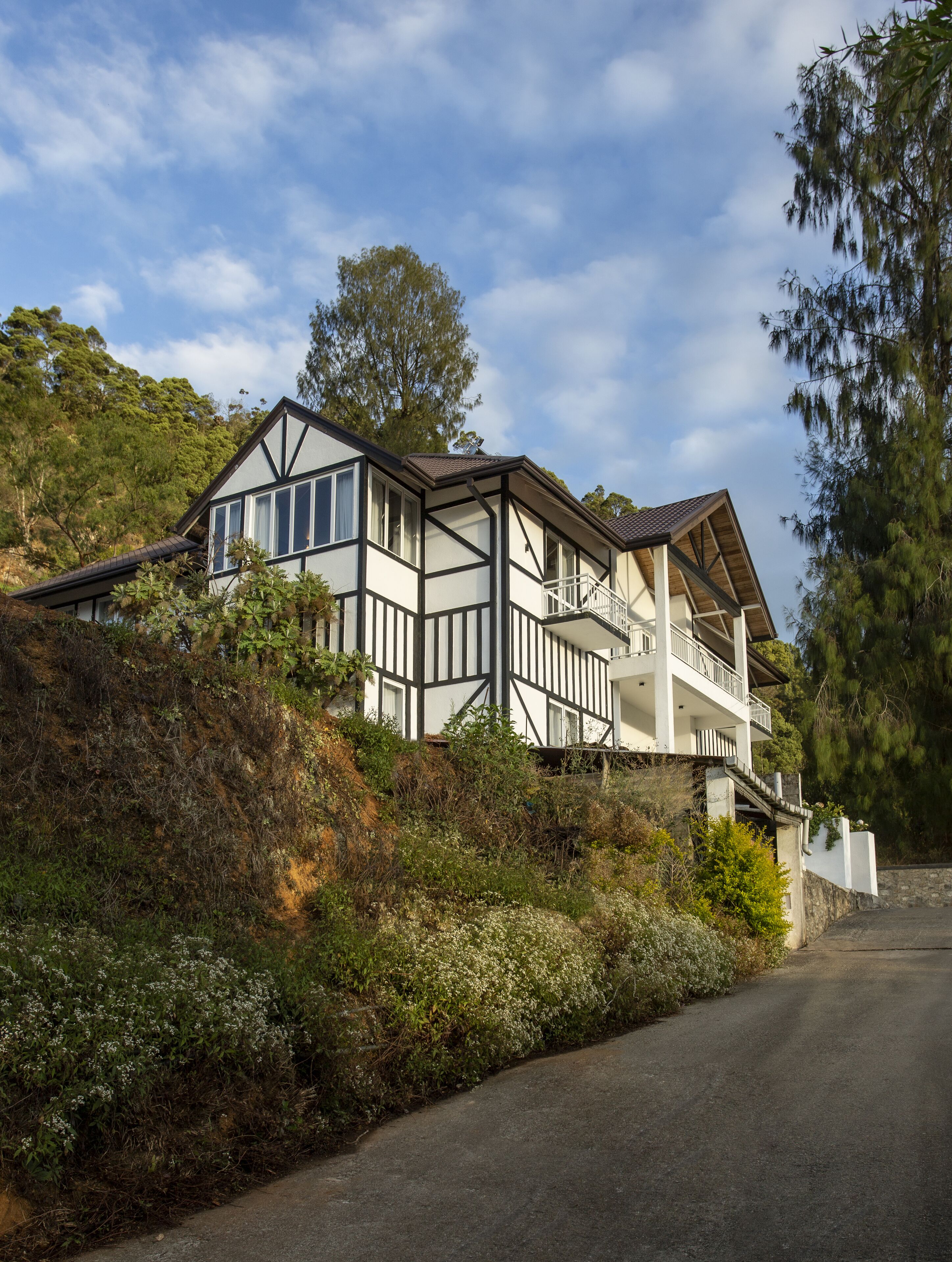 A two-story house with a prominent balcony, situated on a hillside with dense vegetation.