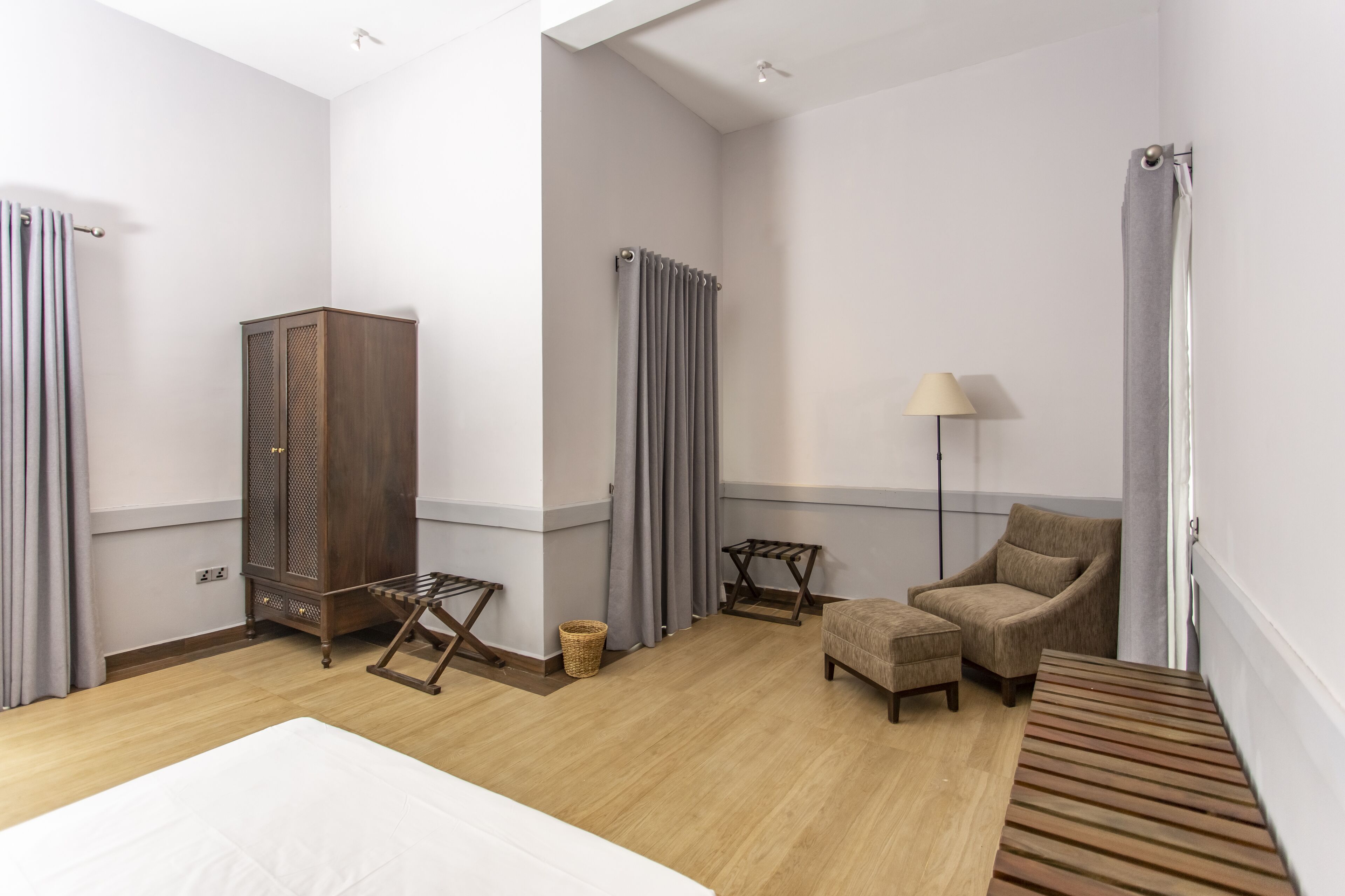A hotel room with a bed, a wooden bench, a chair, a lamp, a wardrobe, and a radiator.