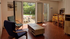 A room with a sliding glass door leading to an outdoor area, a blue chair, a wooden table, and a bookshelf. - Charming Villa: Walk to Beach, Shops, and Forest Retreat in Seignosse (Seignosse)