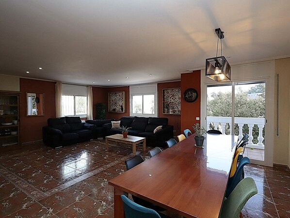 A spacious living room with a dark-colored sofa set, a wooden coffee table, and a dining table with chairs. There are large windows providing natural light and a view of the outdoors.