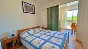 A bedroom with a bed, a nightstand, a lamp, a small table with books, a framed picture on the wall, and a view of a balcony with a chair. - Cozy T2 Apartment with Parking, Pool, and City View Near Beach in Argelès-sur-Mer (Argelès-sur-Mer)