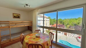 A room with a bunk bed, a dining table with chairs, a large window with a view of a swimming pool and buildings. - Cozy T2 Apartment with Parking, Pool, and City View Near Beach in Argelès-sur-Mer (Argelès-sur-Mer)