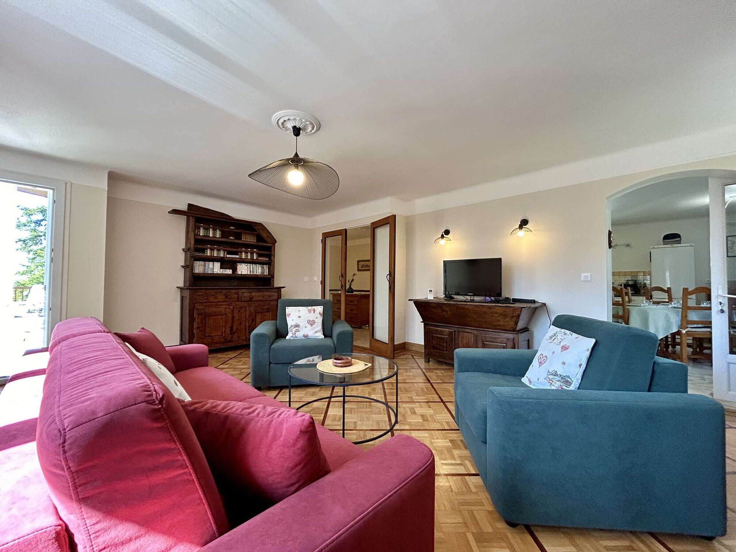 Ein Wohnzimmer mit einem roten Sofa, einem blauen Sofa, einem Holzregal, einem Fernseher und einem Essbereich, der durch eine offene Tür sichtbar ist.