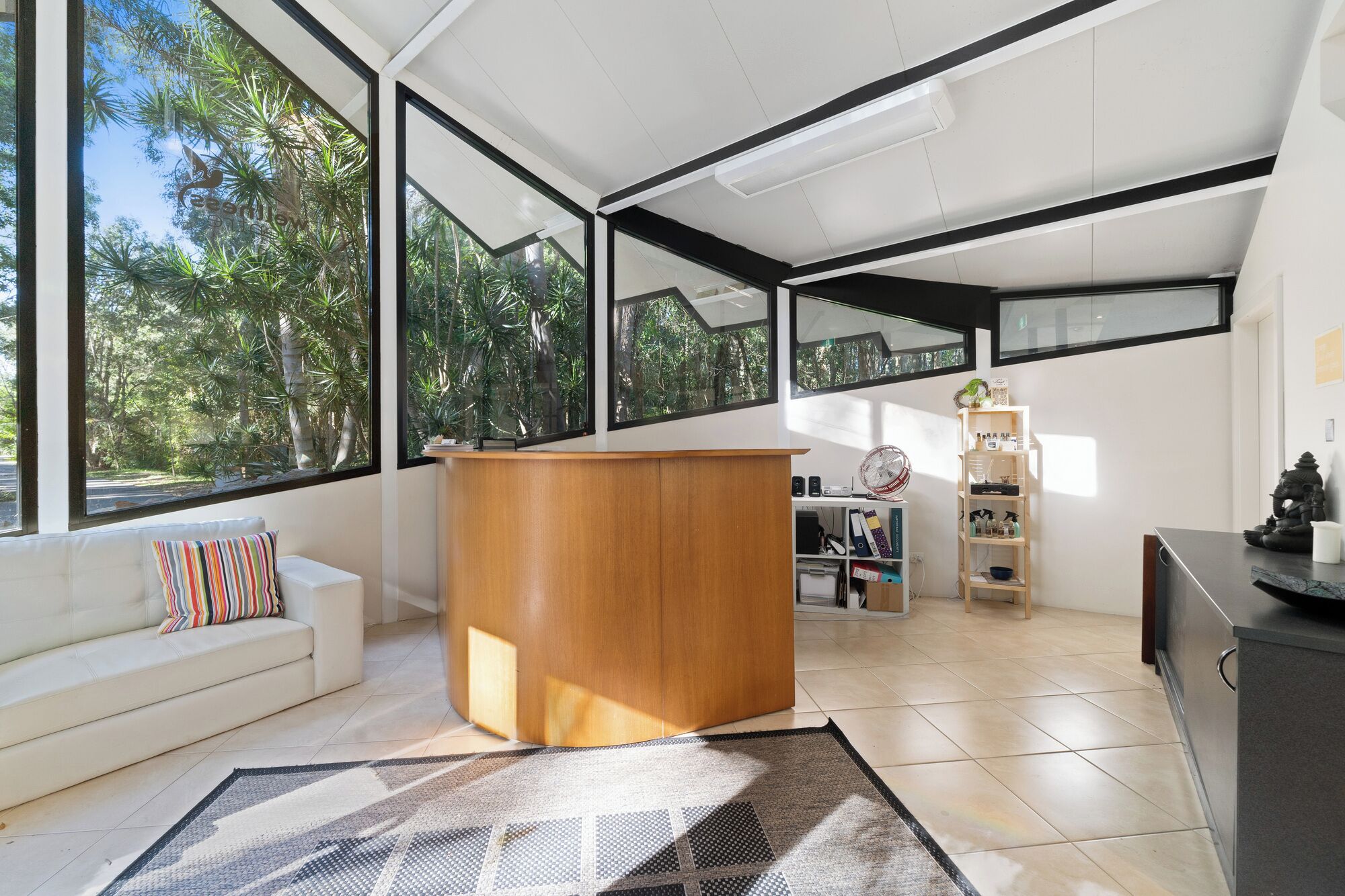A modern living room with a wooden desk, a sofa with colorful pillows, and a view of trees through large windows.