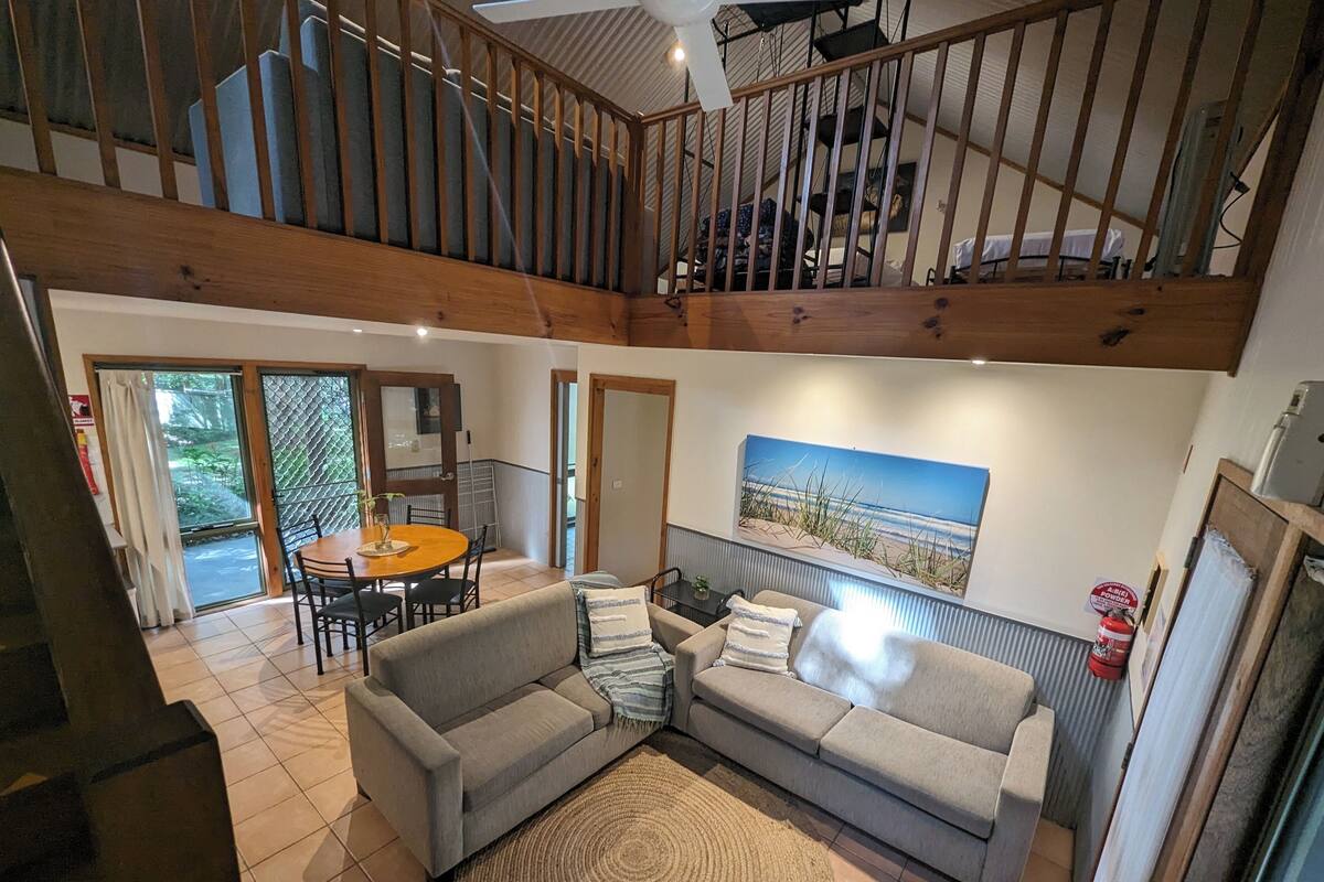 Ein geräumiges Wohnzimmer mit grauem Sofa, einem runden Couchtisch und einem Bild mit einer Strandansicht. Eine Holztreppe führt zu einer oberen Ebene.