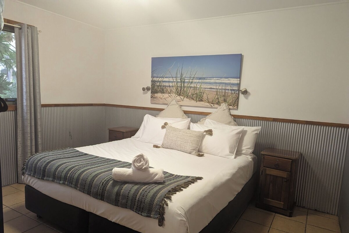 A hotel room with a large bed, a painting of a beach scene, and a window with curtains.