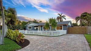 A house with a paved driveway, a white picket fence, and palm trees.