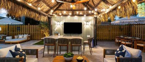 An outdoor patio with a thatched roof, wooden furniture, and a flat-screen TV.