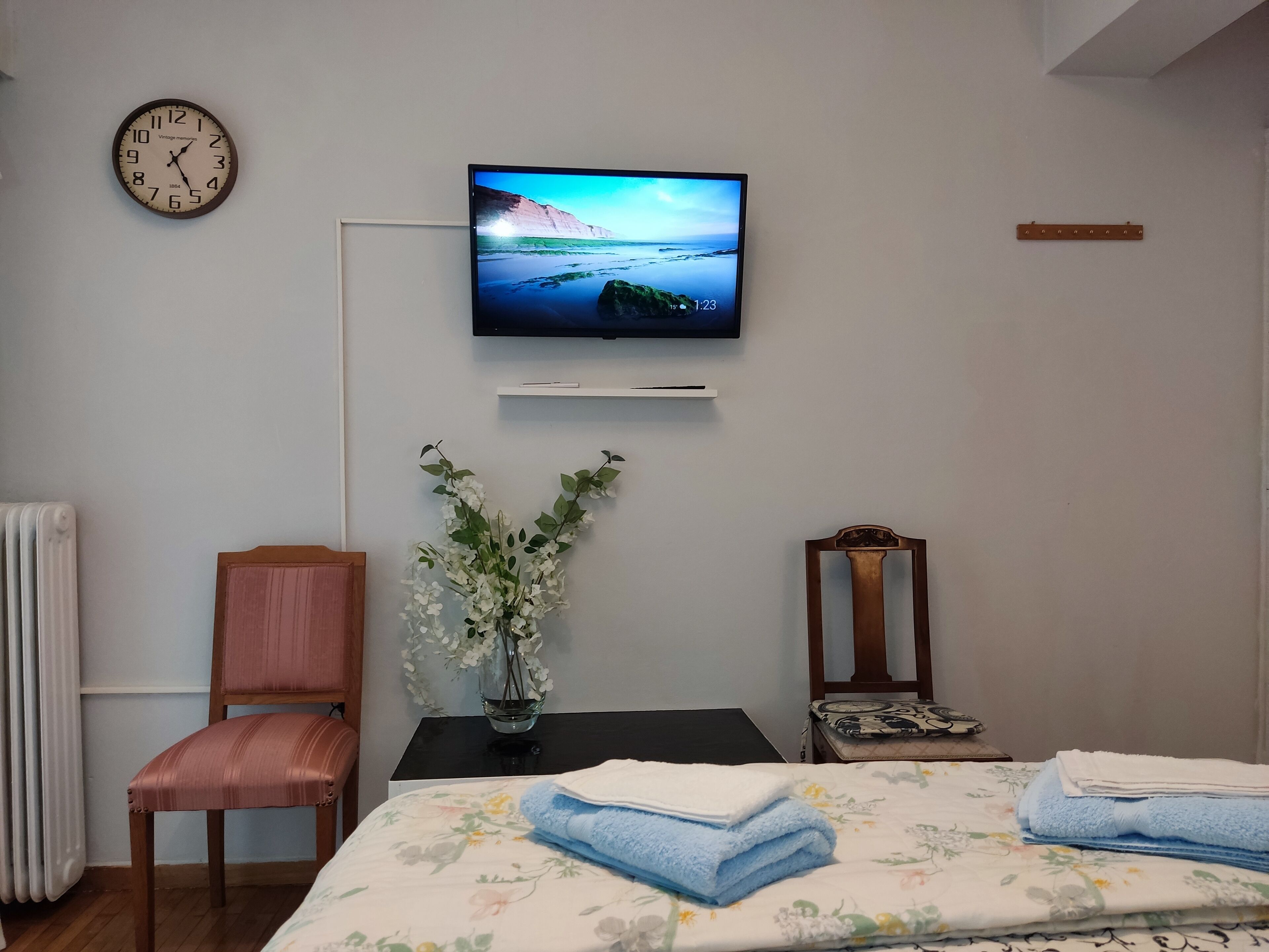 Un dormitorio con cama, una silla, un televisor en la pared, un reloj y un jarrón con flores.